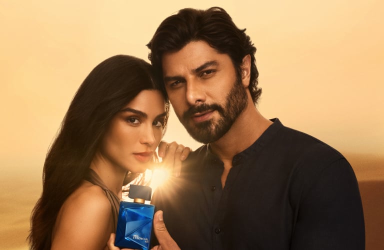 Foto com fundo dourado de deserto. Homem e mulher juntos, enquanto ele segura um frasco azul do perfume Essencial Oud iluminado.